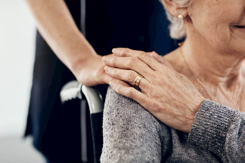 someone placing a hand on an elderly woman's shoulder that is sitting in a wheelchair